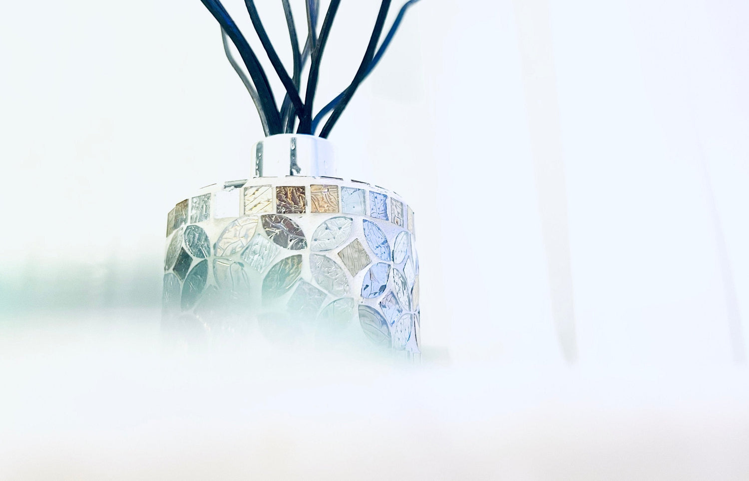 The reed diffuser features a rounded bottle that holds 4 ounces of liquid. The bottle is decorated with a floral mosaic pattern in earthy colors, giving it a natural and artistic look. Its surface feels sandy and has a glass-like finish, adding a unique texture. On top of the bottle, there’s a sleek silver lid. From this lid, several dark reeds rise gracefully. These reeds are curved gently, reminiscent of soft waves, and they enhance the diffuser's elegance and height.